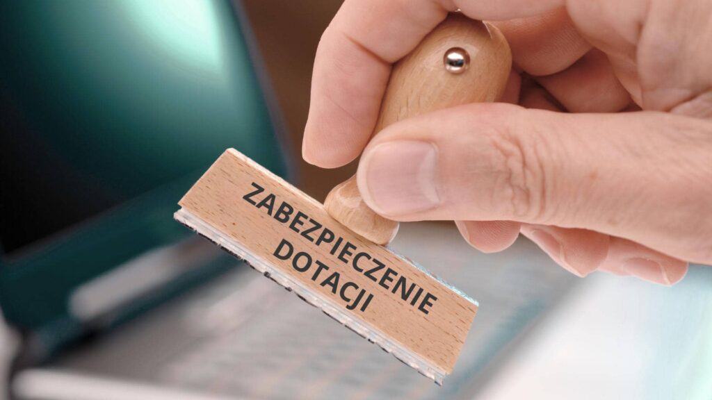 Dłoń na tle laptopa trzymająca piaczątke z napisem ZABEZPIECZENIE DOTACJI.